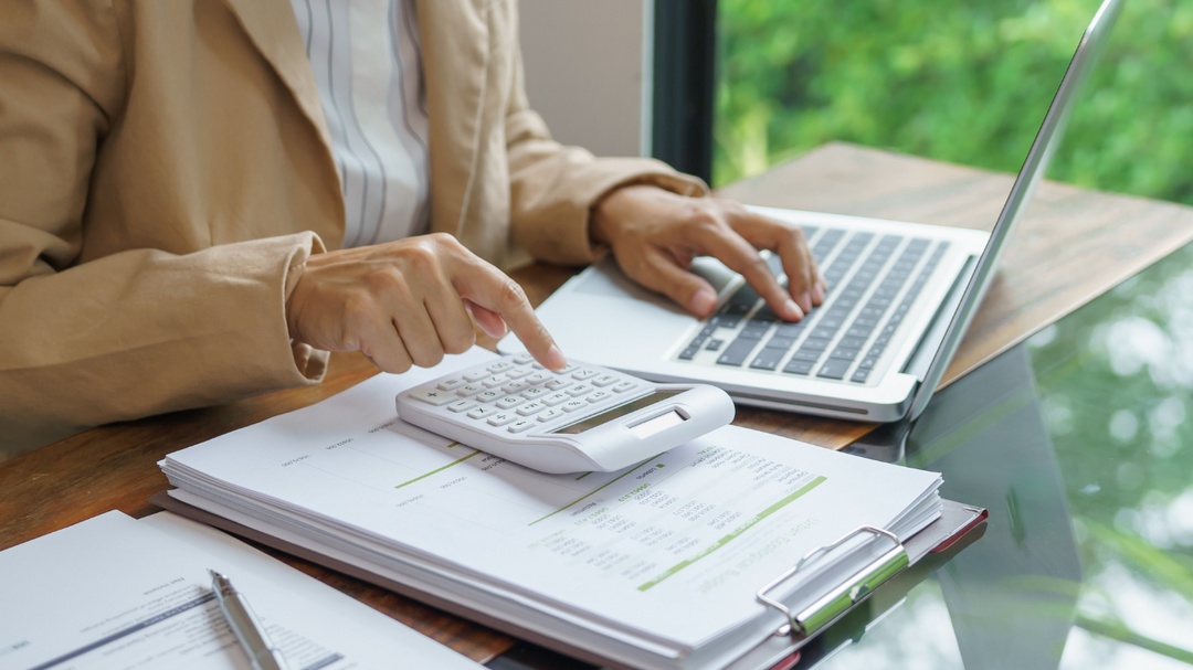 You can see a woman sitting at a desk. You can not see her head. you can only see her from her shoulders down. You would be looking at her from the right of her. she is wearing a tan long sleeve jacket with a white and blue striped shirt underneath. she has her left hand on top of an open tablet keyboard her thumb is on the spacebar and her four other fingers are resting on the keys above the spacebar. her right hand into a fist with index finger pointing at the calculator. There is a stack of papers with a binder clipped to them underneath the calculator. the laptop is to the left side of the calculator and then on the right side there is another stack of papers with a silver pen laying on top of it. the desk underneath the paperwork and laptop is half dark wood and half glass. Behind her is a wall half of it is a window. Outside of the window is blurred green shrubs but bright like the sun is out.